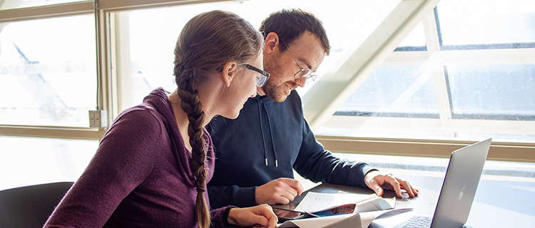 Two person workin together, looking at some documents with a computer in front of them